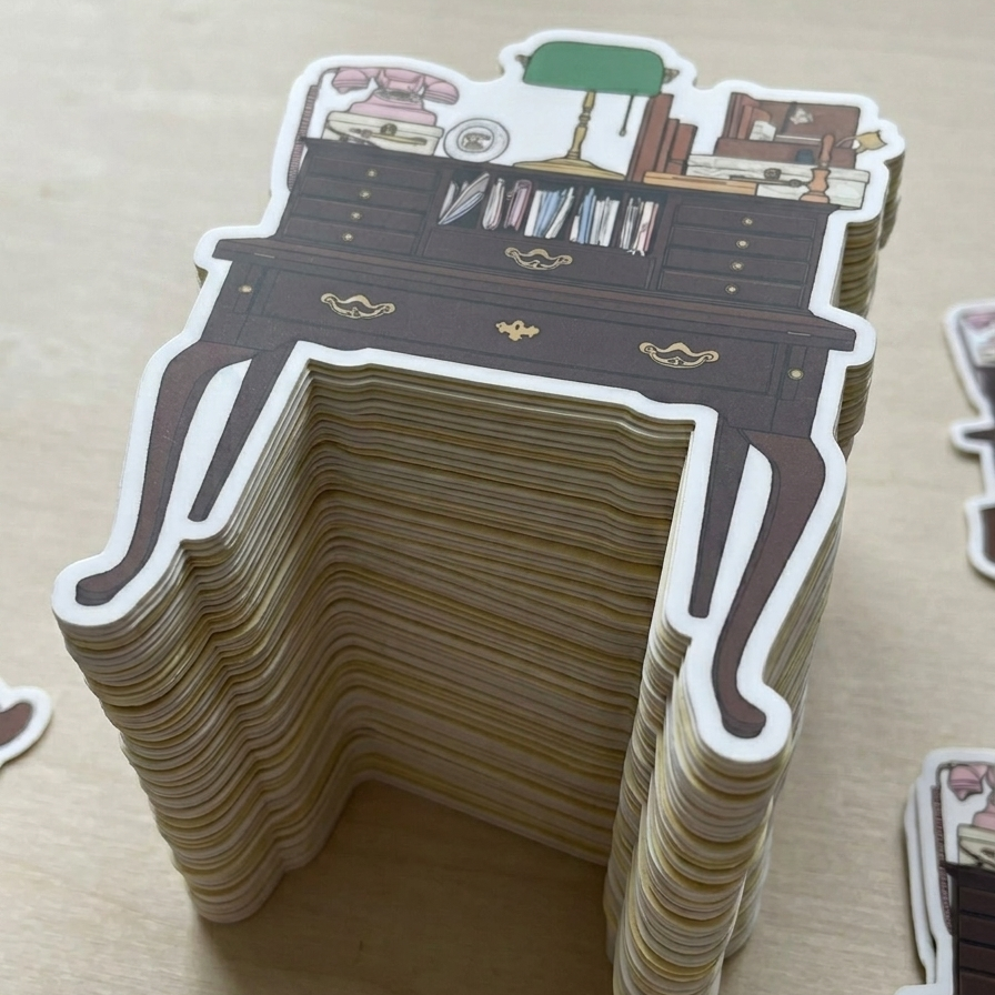 A large stack of die-cut stickers in the shape of a vintage writing desk, illustrated with drawers, gold hardware, journals and notebooks, a pink, vintage-style rotary telephone, and a green banker's lamp, with additional stickers visible at the edges of the frame.