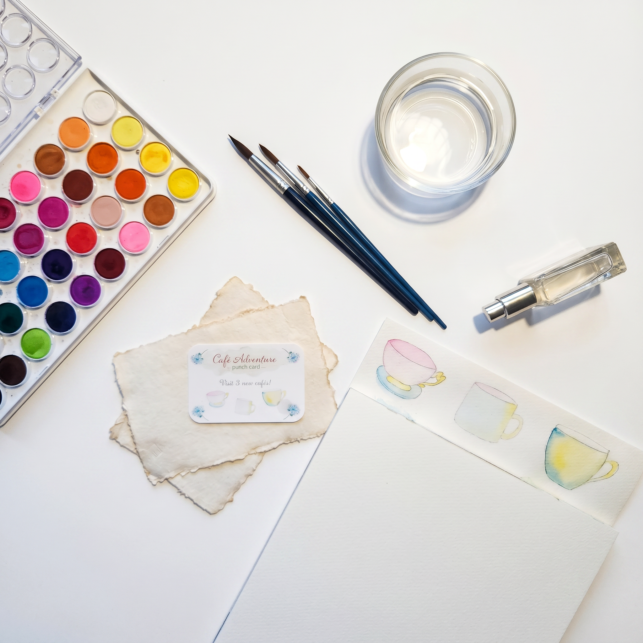 Overhead flat lay of watercolor painting supplies including an open paint palette, three blue-handled brushes, a glass jar with water, and a pump spray bottle, arranged around a Café Adventure punch card resting on torn deckled-edge paper, alongside a partially visible watercolor painting of teacups on white a watercolor pad.